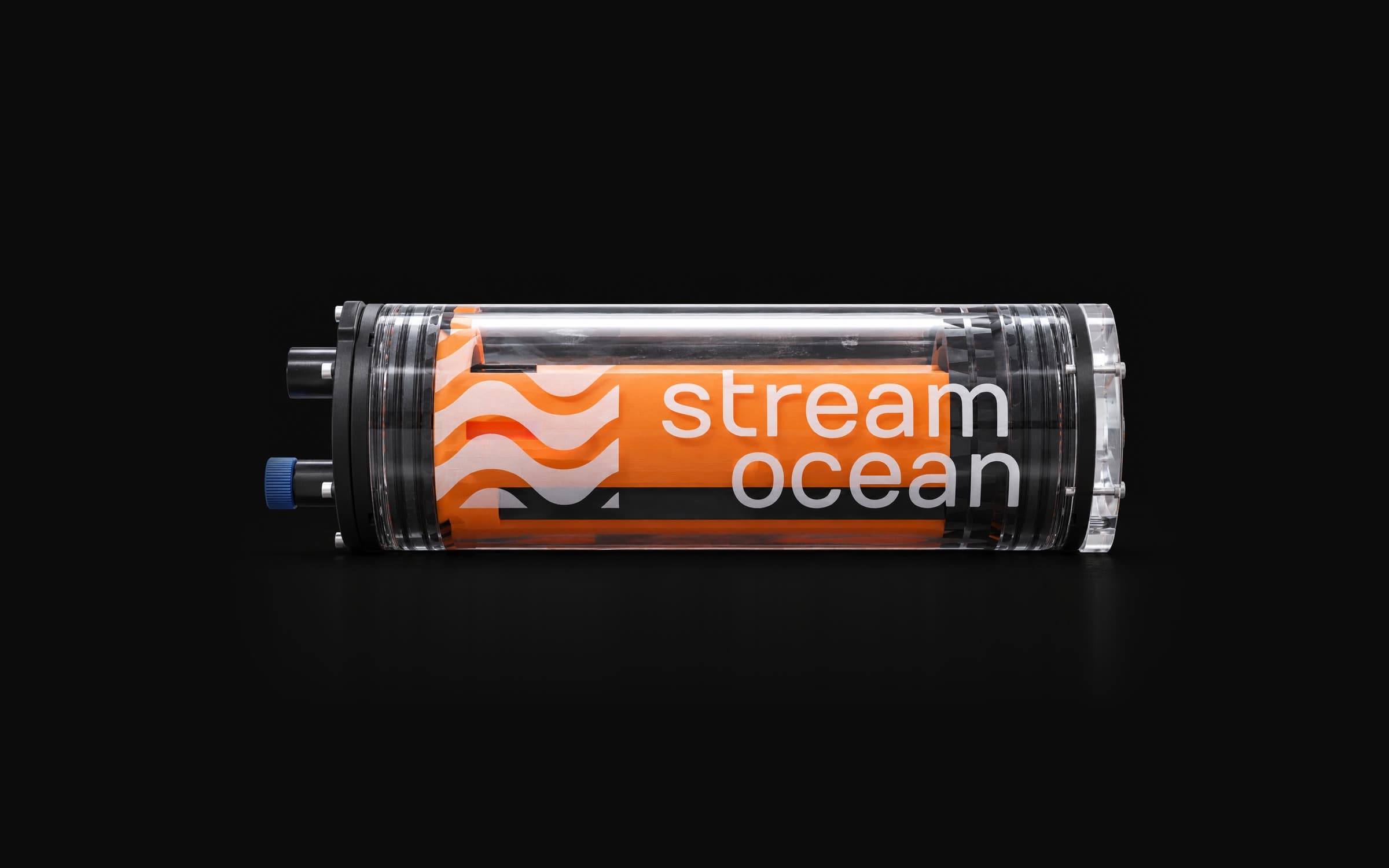 The Stream Ocean camera lying on its side against black: a clear acrylic tube with an orange sleeve inside printed with the white wave mark and stream ocean, black end caps, two connectors on the left, one with a blue cap.