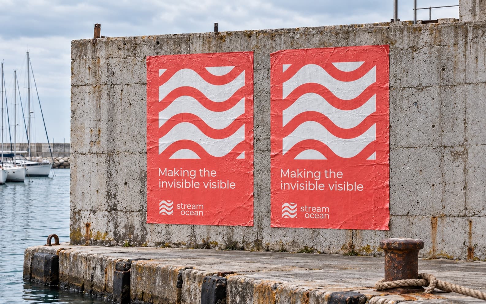 Two Living Coral posters pasted side by side on a weathered concrete harbour wall above a stone quay, a rusty bollard and rope in front and sailing boats on the water to the left: each has the white wave mark large at the top, Making the invisible visible below it, and the stream ocean lockup at the foot.